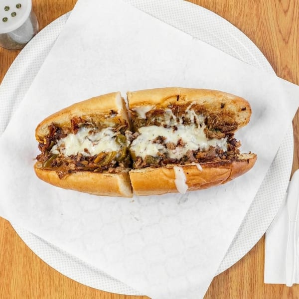 A sliced Philly cheesesteak sandwich with melted cheese, steak, and peppers on a white paper-lined plate.