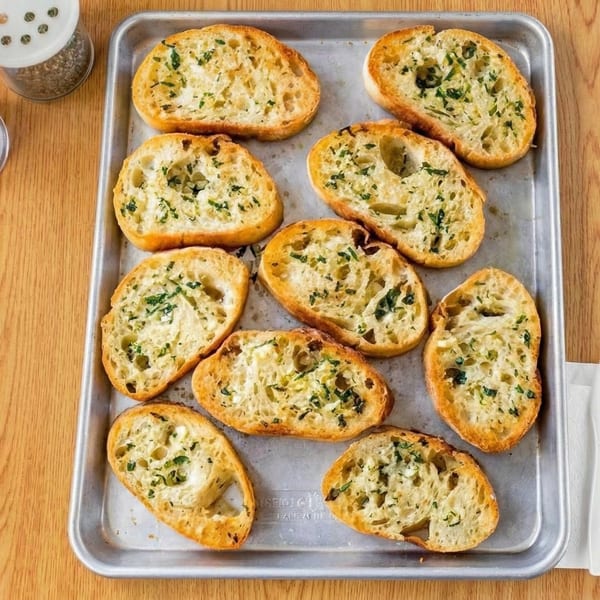 Golden-brown garlic bread slices with green herbs on a metal baking sheet.